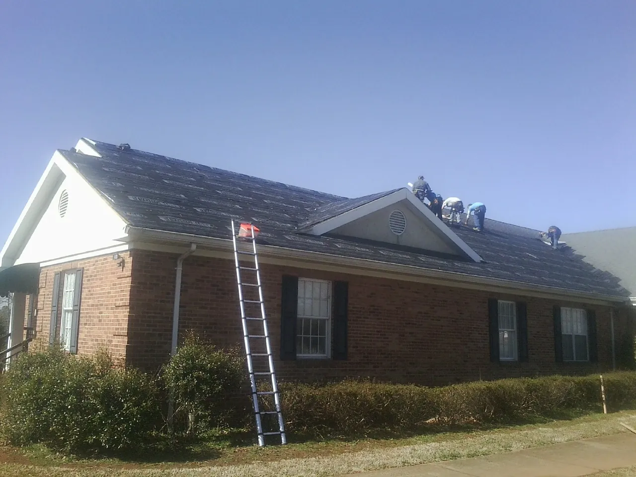 Crew tearing shingles off a brick ranch roof with a ladder set — Emergency Roof Repair work by our crew in Fort Ann