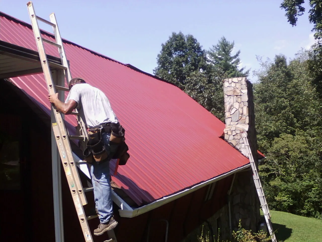 Roofer on a ladder beside a red metal roof and stone chimney — Soffit & Fascia Repair by Roofing Brothers in Fort Ann